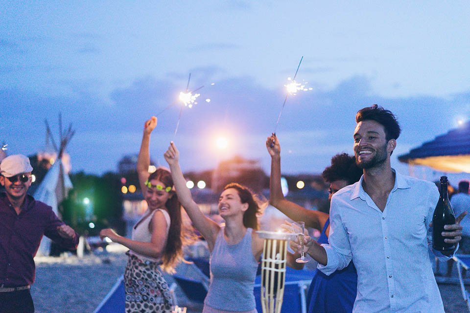 Fröhliche Freunde veranstalten abends eine Strandparty im Freien mit Feuerwerk und trinken Champagner.